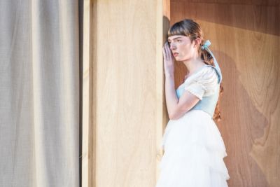 Helena (played by Mary Malone) leans against the wall as she listens in on a conversation happening out of frame. This is a production of “A Midsummer Night’s Dream” performed at Regent’s Park Open Air Theatre (London) in 2026. Photography by Marc Brenner.