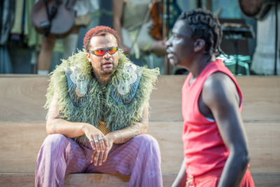 Oberon (played by Olivier Huband) wears a pair of sun glasses and stares at Demetrius (played by Terique Jarrett) in fascination. This is a production of “A Midsummer Night’s Dream” performed at Regent’s Park Open Air Theatre (London) in 2026. Photography by Marc Brenner.