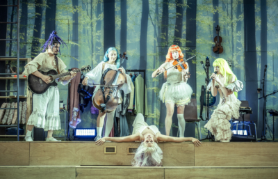 A group of fairies holding various instruments hover over the sleeping Queen of Fairies, Titania (played by Jenny Rainsford). This is a production of “A Midsummer Night’s Dream” performed at Regent’s Park Open Air Theatre (London) in 2026. Photography by Marc Brenner.