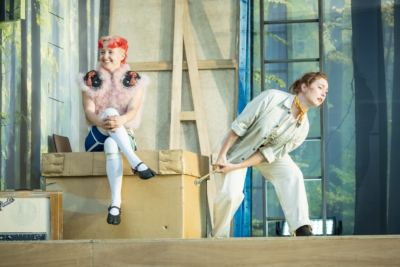 The mischievous fairy Puck (played by G Bruce) sits with their legs crossed on a box that Peter Quince (played by Harriet Anderson-Gordon) is trying to move. Quince doesn’t see Puck and Puck is delighting in their mischief. This is a production of “A Midsummer Night’s Dream” performed at Regent’s Park Open Air Theatre (London) in 2026. Photography by Marc Brenner.