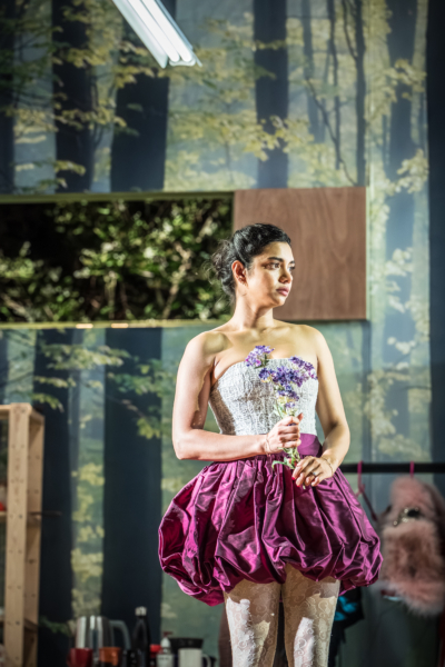 Hermia (played by Hiftu Quasem) wears a purple skirt and holds a purple flower. Behind her, a set backdrop depicting a forest. This is a production of “A Midsummer Night’s Dream” performed at Regent’s Park Open Air Theatre (London) in 2026. Photography by Marc Brenner.