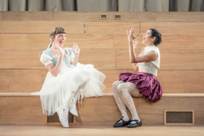 Two women shriek as one of them holds up a ring on their finger. These are Helena (played by Mary Malone) and Hermia (played by Hiftu Quasem). This is a production of “A Midsummer Night’s Dream” performed at Regent’s Park Open Air Theatre (London) in 2026. Photography by Marc Brenner.