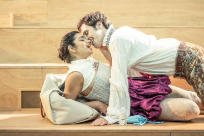 A man straddles over a woman and they both have a passionate look on their faces. They are Lysander (played by Misia Butler) and Hermia (played by Hiftu Quasem). This is a production of “A Midsummer Night’s Dream” performed at Regent’s Park Open Air Theatre (London) in 2026. Photography by Marc Brenner.