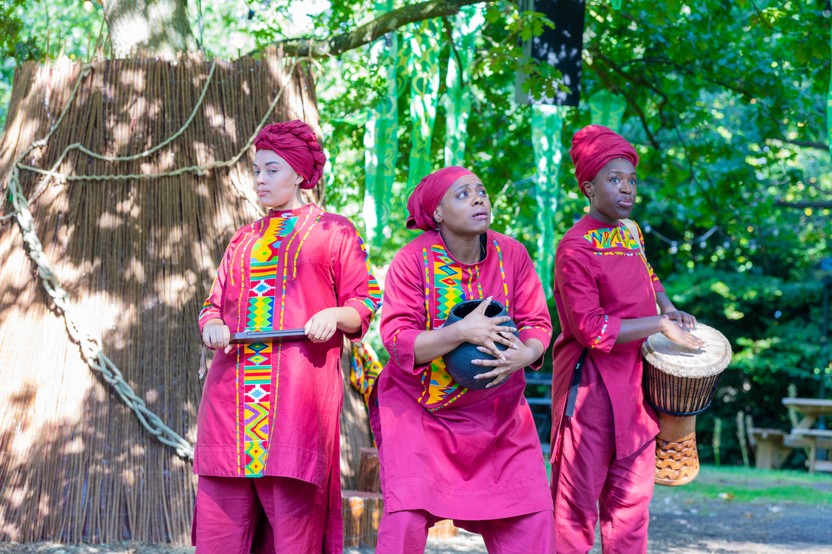 Three women in head scarfs and traditional clothing huddle under a tree. Two of them holds musical instruments whilst the third holds a pot.