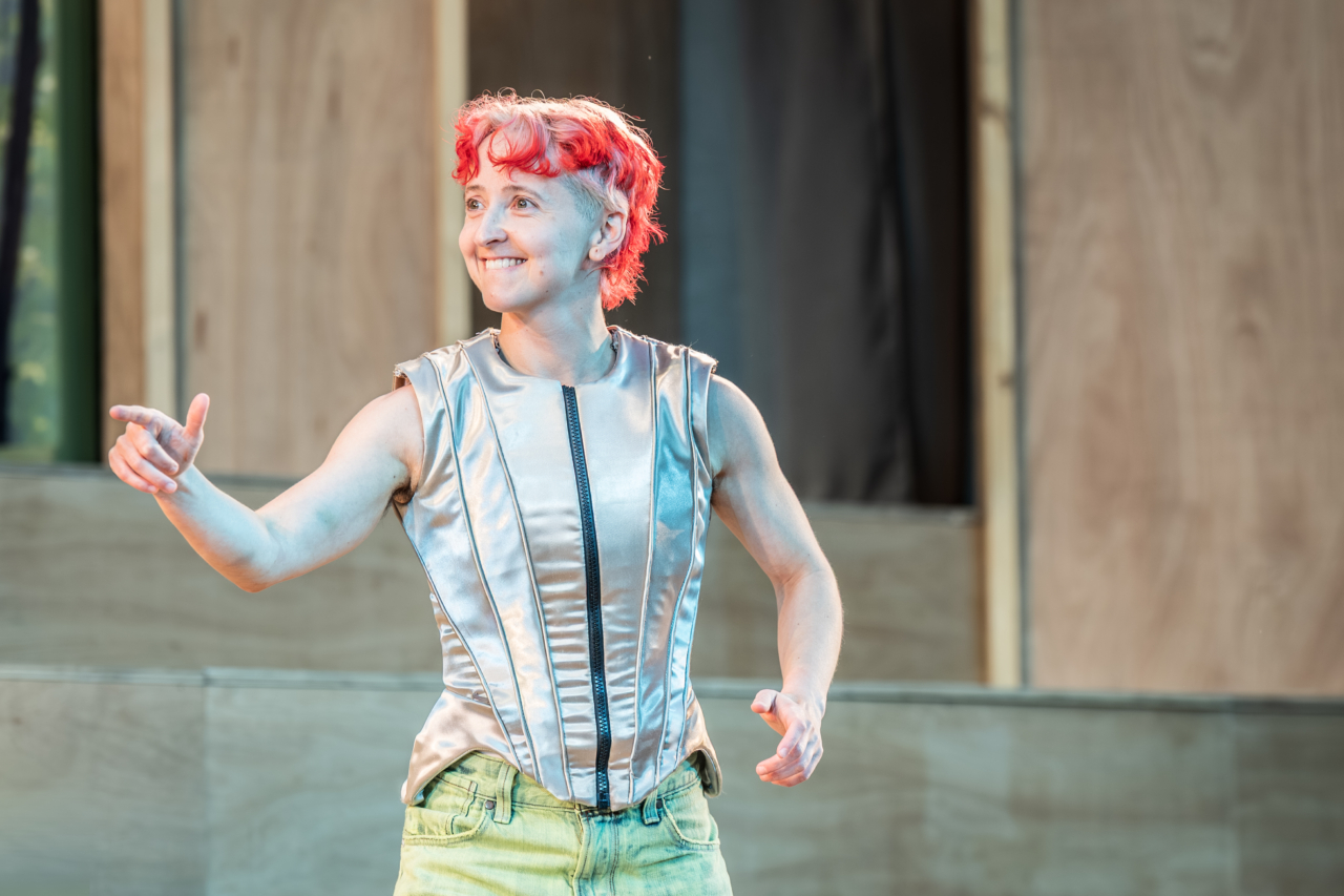 Puck (played by G Bruce) grins cheekily. This is a production of “A Midsummer Night’s Dream” performed at Regent’s Park Open Air Theatre (London) in 2026. Photography by Marc Brenner.