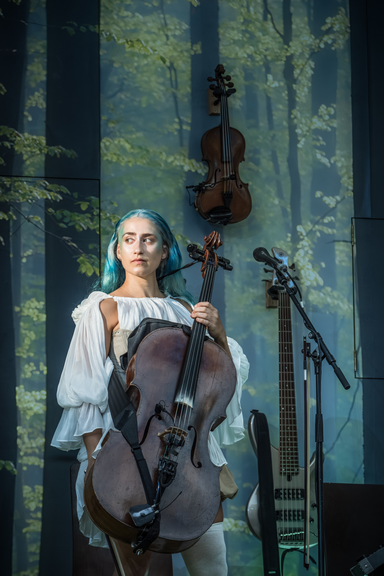 A fairy with blue hair holds a cello. This is a production of “A Midsummer Night’s Dream” performed at Regent’s Park Open Air Theatre (London) in 2026. Photography by Marc Brenner.