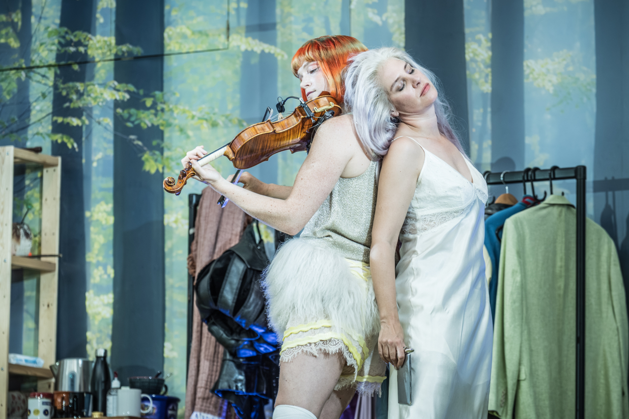 Titania (played by Jenny Rainsford) stands back-to-back with another fairy. Her head lolls back onto the fairy’s shoulder as the fairy plays the violin. This is a production of “A Midsummer Night’s Dream” performed at Regent’s Park Open Air Theatre (London) in 2026. Photography by Marc Brenner.