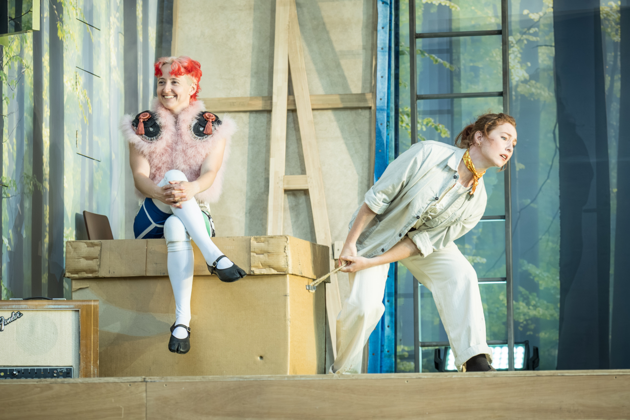 The mischievous fairy Puck (played by G Bruce) sits with their legs crossed on a box that Peter Quince (played by Harriet Anderson-Gordon) is trying to move. Quince doesn’t see Puck and Puck is delighting in their mischief. This is a production of “A Midsummer Night’s Dream” performed at Regent’s Park Open Air Theatre (London) in 2026. Photography by Marc Brenner.