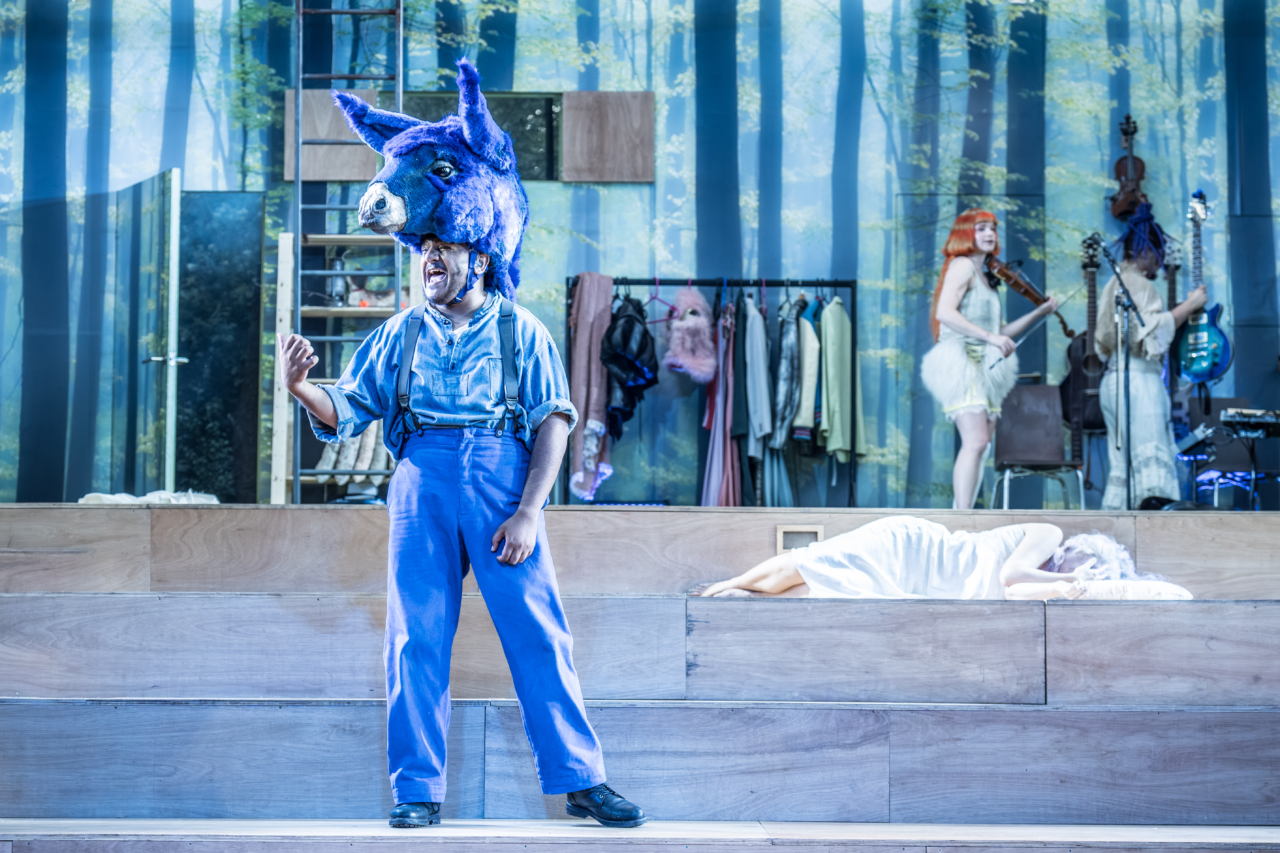 Bottom (played by Nadeem Islam) has a donkey’s head on his head. Bottom and Titania (played by Jenny Rainsford) are sleeping in each others This is a production of “A Midsummer Night’s Dream” performed at Regent’s Park Open Air Theatre (London) in 2026. Photography by Marc Brenner.