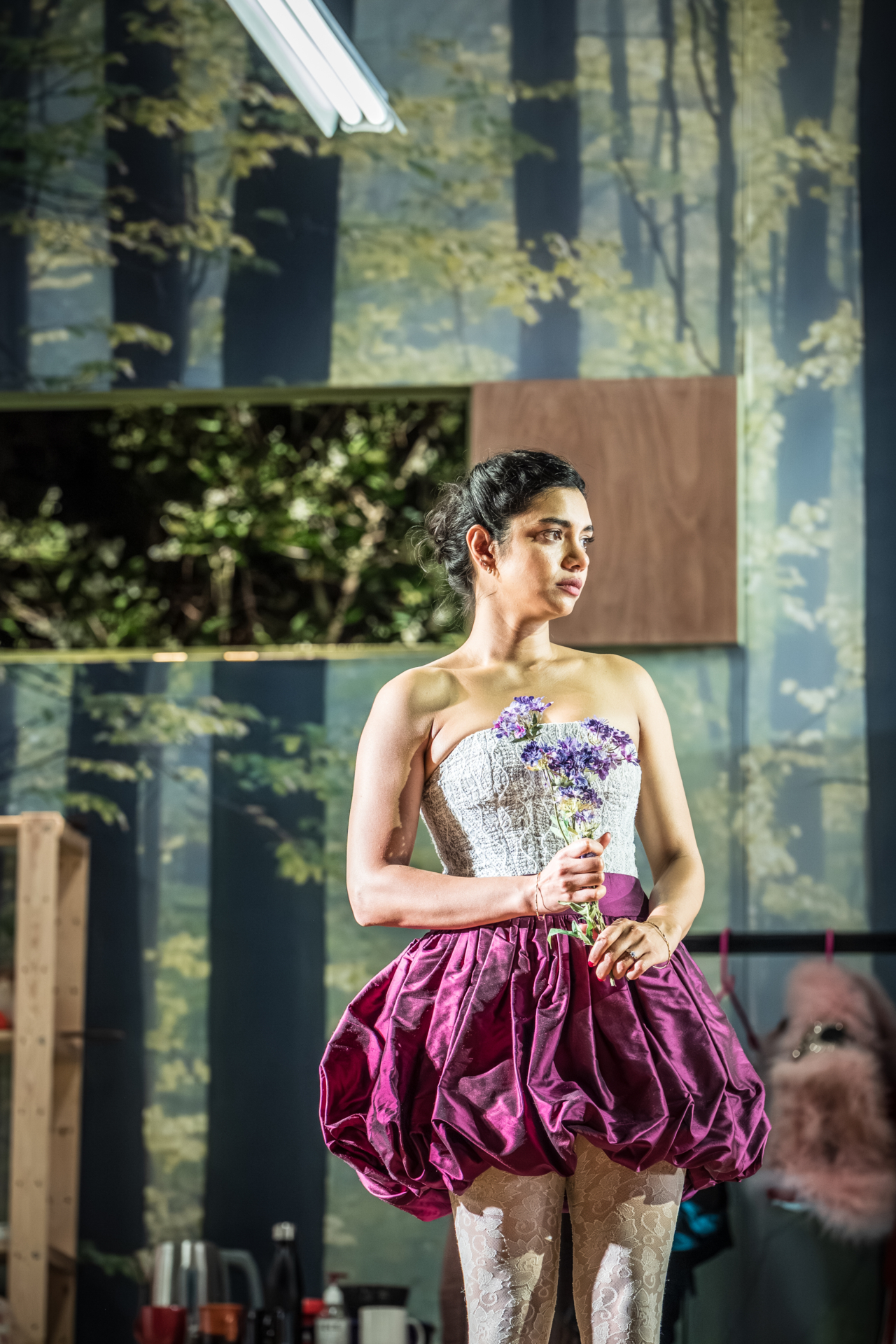 Hermia (played by Hiftu Quasem) wears a purple skirt and holds a purple flower. Behind her, a set backdrop depicting a forest. This is a production of “A Midsummer Night’s Dream” performed at Regent’s Park Open Air Theatre (London) in 2026. Photography by Marc Brenner.