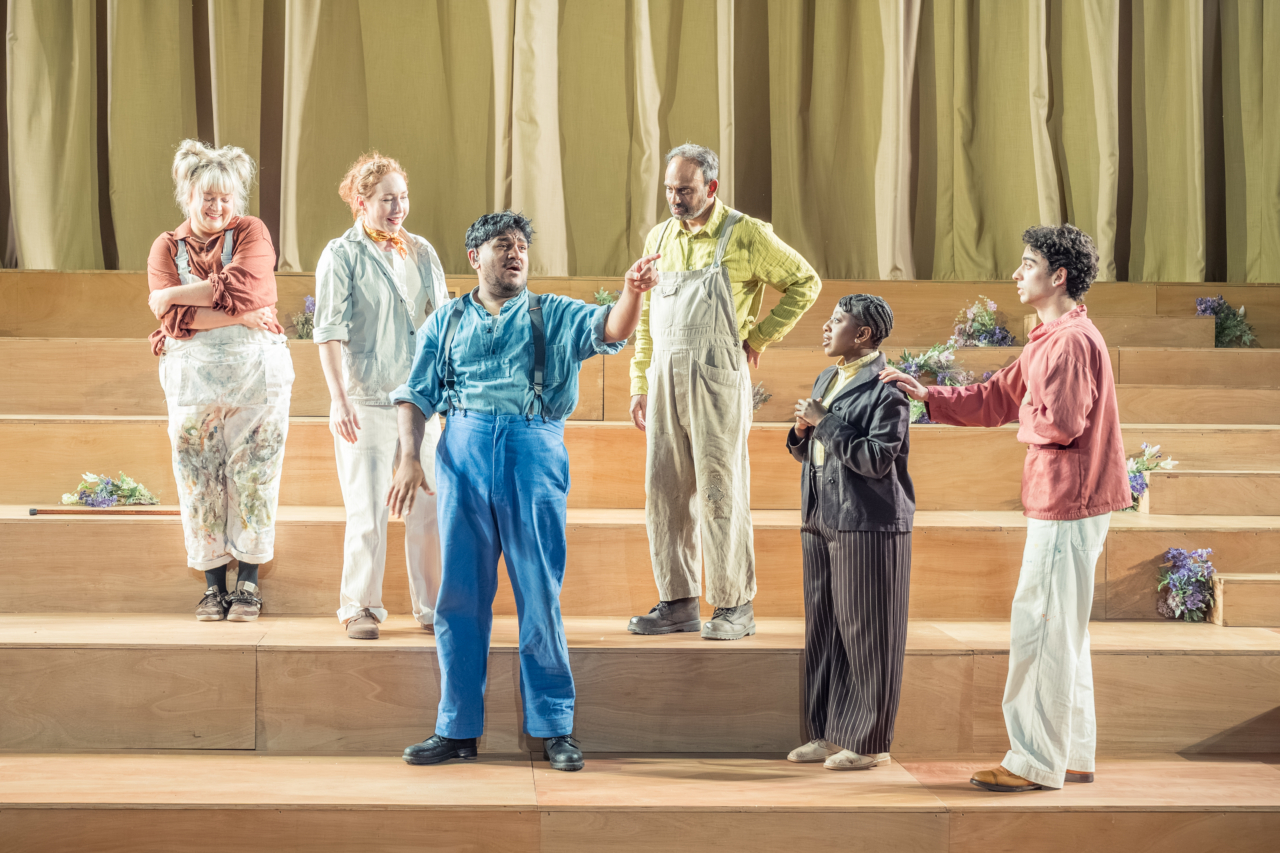 A group of people crowd around a man in blue, who is giving instructions. These are the mechanicals who are crowded around Bottom (played by Nadeem Islam). This is a production of “A Midsummer Night’s Dream” performed at Regent’s Park Open Air Theatre (London) in 2026. Photography by Marc Brenner.