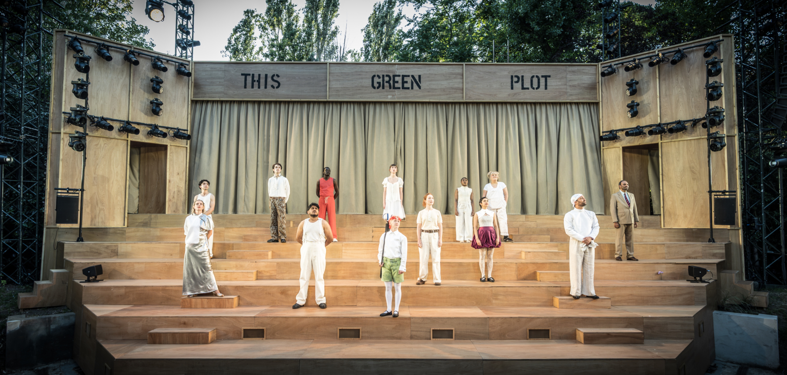 A large wooden stage with curtains and a set of stairs leading down it. On the stairs, the cast of A Midsummer Night’s Dream stands, looking out at the audience. This is a production of “A Midsummer Night’s Dream” performed at Regent’s Park Open Air Theatre (London) in 2026. Photography by Marc Brenner.